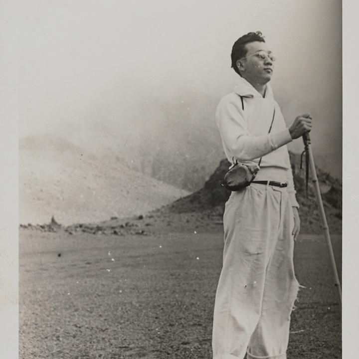Black and white snapshot of Reuben Tam standing with hiking stick and canteen in front of mountains.